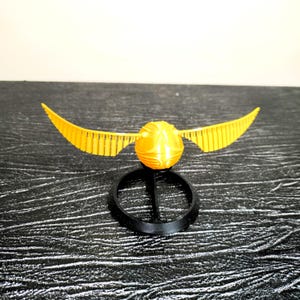 May include: A golden Snitch from the Harry Potter series, with outstretched wings, on a black stand.