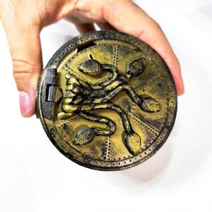Peut inclure: Un récipient rond de couleur or, de style antique, avec un couvercle à charnière. Le couvercle présente un motif en relief détaillé d'une créature ressemblant à un serpent. Le récipient est tenu par une main sur fond blanc.