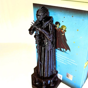 Peut inclure: Un marque-page noir imprimé en 3D représentant une figurine détaillée d'un personnage de roman fantastique. Le marque-page est placé à côté d'un livre avec une couverture bleue et des illustrations de personnages. La figurine est sombre et repose sur une base hexagonale.