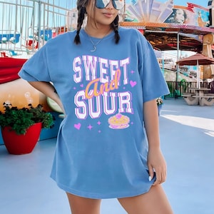 May include: A blue oversized t-shirt with the text 'SWEET and SOUR' in pink and white lettering. The shirt also has a pink heart and a small image of a pie with a cherry on top.