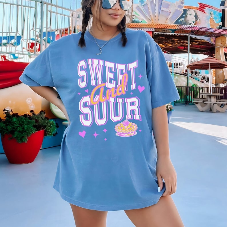 May include: A blue oversized t-shirt with the text 'SWEET and SOUR' in pink and white lettering. The shirt also has a pink heart and a small image of a pie with a cherry on top.