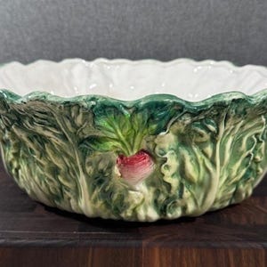 May include: A white ceramic bowl with a green and white cabbage design. The bowl has a raised rim and a small red radish in the center.