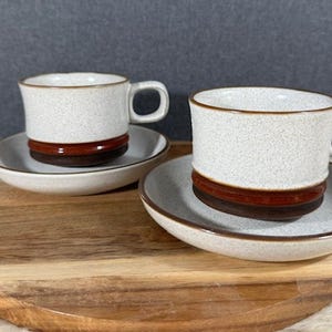 May include: Two white ceramic coffee cups with brown and red bands on the rim, sitting on matching saucers on a wooden surface.