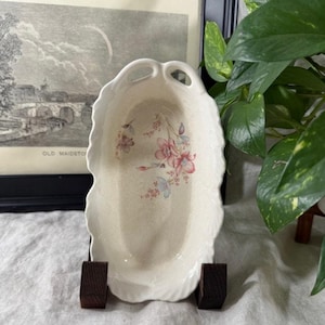 Puede incluir: Plato de cerámica ovalado de color crema, con borde festoneado y dos asas de bucle. El plato presenta un estampado floral en rosa y azul. Se exhibe sobre un soporte de madera oscura, con una impresión en blanco y negro enmarcada en el fondo.