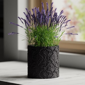 May include: A potted lavender plant with vibrant purple flowers and green foliage. The plant is in a black, cylindrical pot with an embossed floral pattern. The pot sits on a white surface near a window.