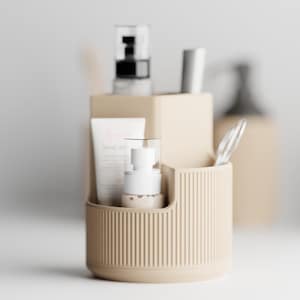 Puede incluir: Un organizador cilíndrico beige y acanalado que contiene productos para el cuidado de la piel. El organizador contiene un tubo de loción, un frasco con bomba blanco y otros recipientes cosméticos. El fondo presenta recipientes beige adicionales y un fondo blanco borroso.