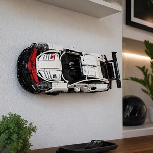 May include: A white, black, and red Porsche 911 RSR model car constructed from building blocks, mounted horizontally on a wall. The vehicle features visible headlights, a rear spoiler with Porsche branding, and Mobil 1 and 911 decals.