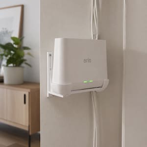 May include: A white plastic wall mount bracket holding an Arlo smart hub device. The hub is white with the Arlo logo printed on the front and two illuminated green status lights. White cables run vertically behind the device along the wall.