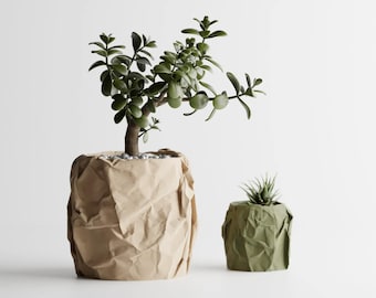 May include: A large jade plant in a tan, crumpled paper-style planter and a smaller succulent in a sage green, crumpled paper-style planter. Both planters have a textured, creased appearance resembling folded paper bags with rounded edges.