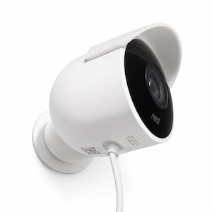 May include: White security camera with a black lens and a protective visor. The camera has the word "nest" printed on the front and is mounted on a white surface. A white cable extends from the camera.