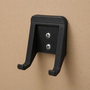Könnte beinhalten: Ein schwarzer Wandhalter mit zwei gebogenen Armen und einer rechteckigen Rückplatte. Zwei silberne Schrauben befestigen den Halter an einer beigefarbenen Wand. Der Halter ist zur Aufnahme eines Gegenstandes bestimmt.
