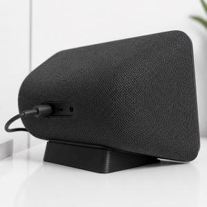 May include: A charcoal gray smart speaker with a fabric-covered exterior and a black plastic base. The back panel features a central recessed area containing a power input port and a small circular auxiliary jack, with a black power cable connected.
