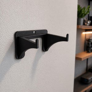 May include: A black wall-mounted shelf bracket with two curved support arms and two hooks. The bracket is attached to a textured, light gray wall with a single screw. The design is minimalist and modern.