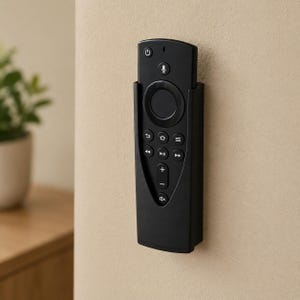 Op de afbeelding: Een zwarte afstandsbediening met meerdere knoppen en een microfoonpictogram is aan een beige muur bevestigd. De afstandsbediening zit in een zwarte houder. Op de achtergrond is een kleine plant te zien.