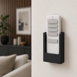 May include: A white remote control with several gray buttons is held in a black wall-mounted holder. The remote has a red light at the top. The holder is rectangular and mounted on a white wall. The remote is for controlling a ceiling fan.