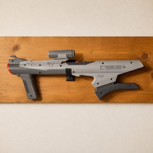 Op de afbeelding: Een grijze Nintendo Scope lichtpistool-controller met een afneembaar vizier, een voorste handgreep en een schoudersteun. Het apparaat heeft een rode punt bij de loop, zwarte accenten op de handgrepen en de tekst Nintendo Scope op de behuizing.