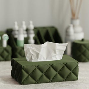 Puede incluir: Un dispensador de pañuelos verde oscuro, acolchado, con un pañuelo blanco que sobresale. El dispensador presenta un patrón de diamantes y forma parte de un conjunto de accesorios de baño.