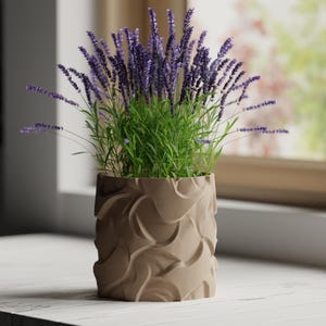 May include: A lavender plant in a beige, textured pot. The plant has purple flowers and green leaves. The pot has a wave-like design. The pot is on a white surface near a window, with a view of the outside.