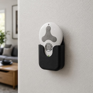 May include: A white remote control with a gray triangular button layout featuring the numbers 1, 2, and 3, and a circular button at the bottom, housed in a black wall-mounted holder attached to a white wall surface.