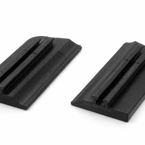 May include: Two identical black rectangular plastic components featuring a rounded edge on one side and a raised central track or groove running along the length of the top surface. These pieces have a flat base and a textured matte finish.