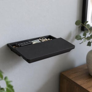 May include: A black wall-mounted shelf with a pull-out drawer. The drawer is partially open, revealing a car key fob, cash, jewellery, and other small items. The shelf is mounted on a light grey wall. A vase with greenery is in the background.