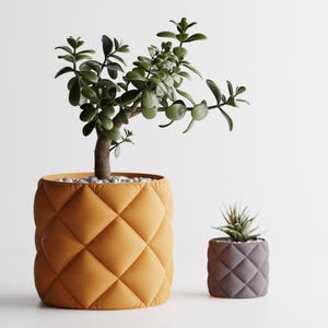 May include: Two potted plants with a quilted design. The larger plant has green leaves and is in a yellow pot. The smaller plant has green leaves and is in a gray pot.