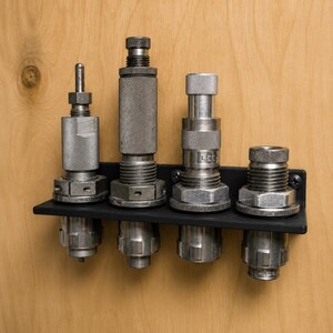 May include: A black metal storage rack holding four silver reloading dies. The rack is mounted horizontally and features a matte finish. Each die has a cylindrical shape with knurled sections, threaded bodies, and hexagonal nuts for secure adjustment.