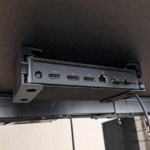 May include: A black under-desk mounting bracket holding a small computer. The device features a power port, an HDMI port, two DisplayPort inputs labeled D1 and D2, an Ethernet port, and two USB ports. The bracket is secured to a flat black surface.
