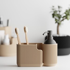Puede incluir: Un juego de organizador de baño beige con un soporte para cepillos de dientes, un dispensador de jabón y una pequeña planta en el fondo. El soporte para cepillos de dientes tiene dos cepillos de dientes. El dispensador de jabón tiene una bomba gris oscuro.