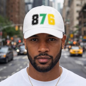 May include: White baseball cap featuring the number "876" in black, green, and yellow. The hat is worn by a person in front of a city street background. The cap is a classic baseball style.