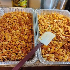 Könnte beinhalten: Zwei Backbleche aus Aluminiumfolie gefüllt mit einer Mischung aus goldbraunen Cerealienquadraten, Salzstangen und anderen Snacks. Ein weißer und brauner Spatel liegt zwischen den Blechen, was darauf hindeutet, dass der Inhalt gemischt oder serviert wird. Der Snack-Mix scheint gewürzt zu sein.