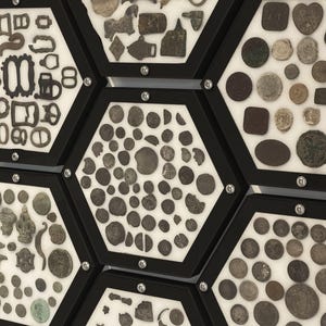 May include: A collection of antique metal artefacts and coins displayed in multiple black hexagonal frames. The items include various buckles, fasteners, and circular coins of different sizes and textures, all mounted on a white background.