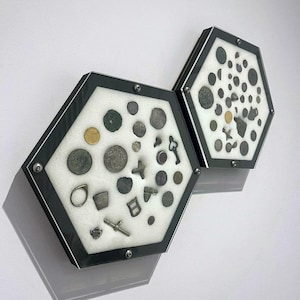 Puede incluir: Dos vitrinas hexagonales con marcos negros e interiores de espuma blanca, que muestran una colección de monedas antiguas, joyas y otros pequeños artefactos.