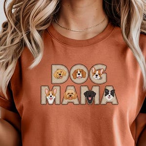 Puede incluir: Camiseta de color óxido con el texto "DOG MAMA" en un diseño cosido. La palabra "DOG" presenta ilustraciones de tres razas de perros diferentes, y "MAMA" presenta ilustraciones de dos razas de perros más. La camiseta tiene mangas enrolladas.