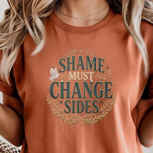 以下が含まれることがあります： 袖をまくった錆色のTシャツ。Tシャツには、「SHAME MUST CHANGE SIDES」の文字が、鳩と花のアクセントをあしらった装飾的なデザインで表示されています。Tシャツはデニムショーツと組み合わせています。