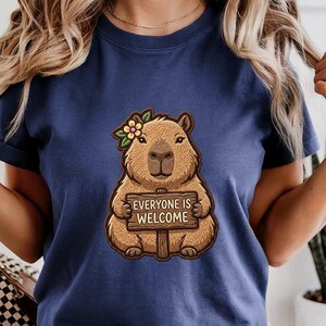 May include: Navy blue t-shirt featuring a cartoon capybara with a flower crown, holding a sign that reads "EVERYONE IS WELCOME." The capybara is brown with a light brown belly. The shirt has rolled sleeves.