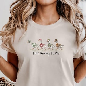 May include: A beige t-shirt featuring a whimsical design of four geese, each adorned with a different patterned hat. The text "Talk Derby To Me" is printed below the geese. The shirt is made of a soft material.