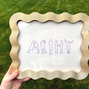 Puede incluir: Un marco de fotos rectangular con un borde ondulado y texturizado en tono dorado. El inserto de vidrio presenta un dise&ntilde;o bordado en color morado que representa las letras de la palabra ARMY, donde cada letra est&aacute; formada por la silueta de una persona.