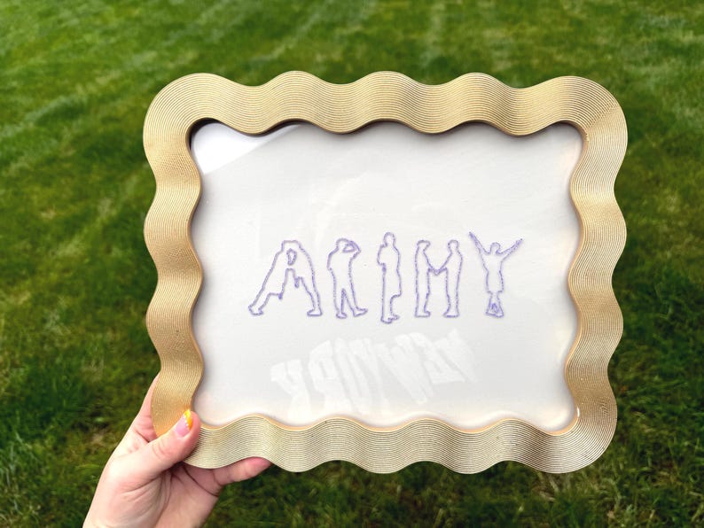 Puede incluir: Un marco de fotos rectangular con un borde ondulado y texturizado en tono dorado. El inserto de vidrio presenta un dise&ntilde;o bordado en color morado que representa las letras de la palabra ARMY, donde cada letra est&aacute; formada por la silueta de una persona.