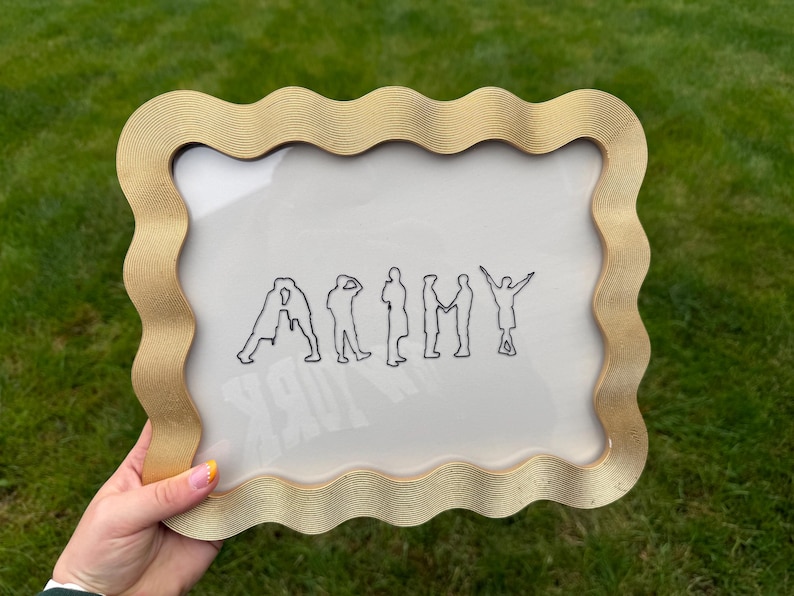 Puede incluir: Un marco de fotos rectangular con un borde dorado ondulado y texturizado. Dentro del marco, un fondo blanco presenta una ilustraci&oacute;n de l&iacute;neas negras de siete figuras dispuestas para deletrear la palabra ARMY, formando cada una una letra.