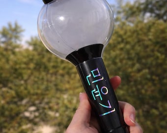 Puede incluir: Un bastón luminoso negro con una cúpula superior esférica transparente y un botón rojo en la parte superior. El mango es negro con texto coreano holográfico iridiscente impreso verticalmente en el lateral. Incluye una correa gris.