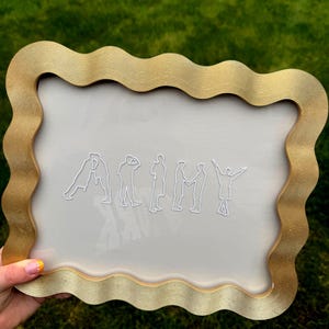 Puede incluir: Un marco rectangular con un borde dorado ondulado que rodea un panel transparente. El centro presenta siluetas de l&iacute;neas blancas de siete figuras dispuestas para deletrear la palabra ARMY, donde cada figura representa una letra.