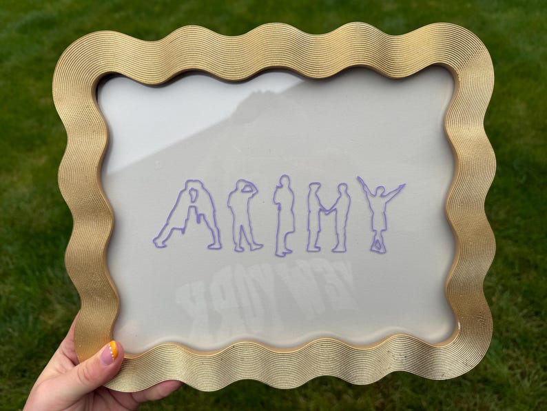 Puede incluir: Un marco rectangular con un borde ondulado y texturizado de color dorado que rodea una pantalla transparente. La pantalla presenta un gr&aacute;fico de l&iacute;neas moradas que deletrea la palabra ARMY, donde cada letra es la silueta de una persona.
