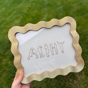 Puede incluir: Un marco de fotos rectangular con un borde dorado ondulado y texturizado. El marco contiene un inserto blanco con una ilustraci&oacute;n de l&iacute;neas negras de siete siluetas humanas dispuestas para deletrear la palabra ARMY, con la &uacute;ltima figura alzando los brazos.