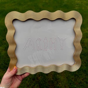 Puede incluir: Un marco rectangular con un borde ondulado de color dorado que presenta una superficie texturizada y estriada. El centro es un panel transparente y claro que muestra siluetas de l&iacute;neas blancas de siete figuras que forman la palabra ARMY.