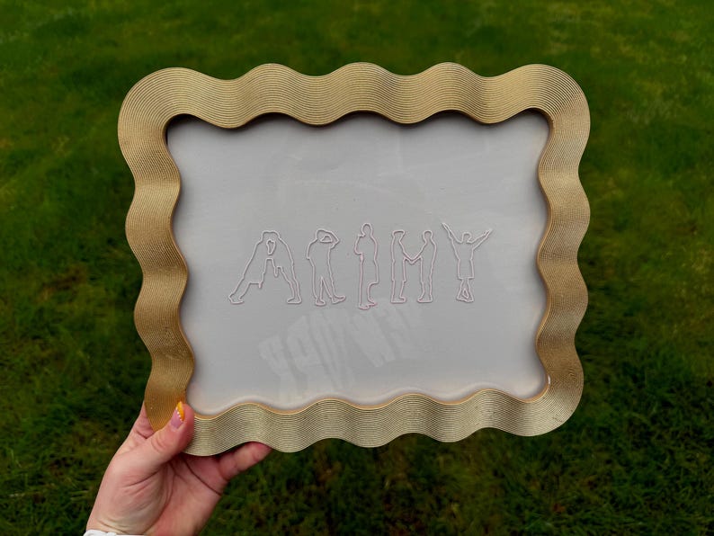 Puede incluir: Un marco rectangular con un borde ondulado de color dorado que presenta una superficie texturizada y estriada. El centro es un panel transparente y claro que muestra siluetas de l&iacute;neas blancas de siete figuras que forman la palabra ARMY.