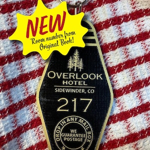 May include: A black and gold hotel keychain with the text "OVERLOOK HOTEL SIDEWINDER, CO 217". A yellow starburst tag reads "NEW Room number from Original Book!". The keychain has a metal ring and the words "DROP IN ANY MAIL BOX WE GUARANTEE POSTAGE".