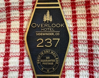 May include: A black and gold hotel key chain with the text "OVERLOOK HOTEL SIDEWINDER, CO" and the number "237". The key chain also has a gold circle with the text "DROP IN ANY MAIL BOX WE GUARANTEE POSTAGE" and three stars.