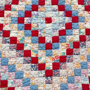 May include: A handmade quilt with a patchwork design. The quilt features a grid pattern of red squares alternating with squares of various floral and patterned fabrics in shades of blue, white, and yellow. The quilt has a textured, quilted surface.