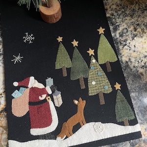 May include: A felt Christmas table runner with a black background, featuring a Santa Claus figure carrying a bag of gifts and a lantern, a fox, and several green Christmas trees with gold stars. The runner has a white snow-covered border.
